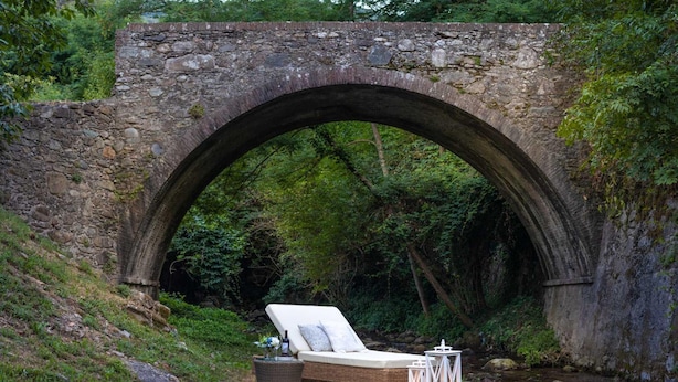 Un puente de piedra sobre un arroyo, con una silla de descanso y un farol debajo.