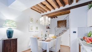 Un comedor con mantel blanco, sillas de madera, una araña y una escalera al fondo.