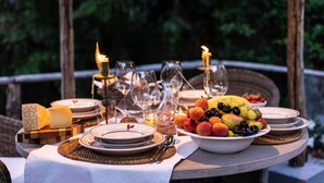 Mesa al aire libre con platos, vasos y frutero, rodeada de velas y una silla de mimbre.