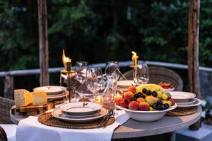 Ein Tisch im Freien, gedeckt mit Tellern, Gläsern und einer Obstschale, umgeben von Kerzen und einem Korbstuhl.