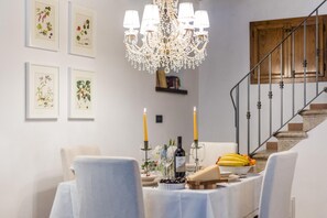 Ein Essbereich mit einer Kristallleuchtre, gerahmten Pflanzenabbildungen und einem Tisch, der mit Kerzen, Wein und Früchten gedeckt ist.