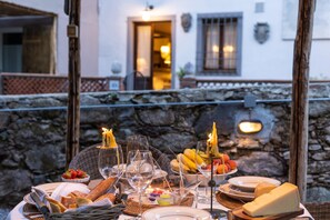 Un espace de restauration en plein air, avec un mur de pierre en arrière-plan, un panier rempli de pain, de fruits et de fromage, et une table agrémentée de verres à vin et d’assiettes.