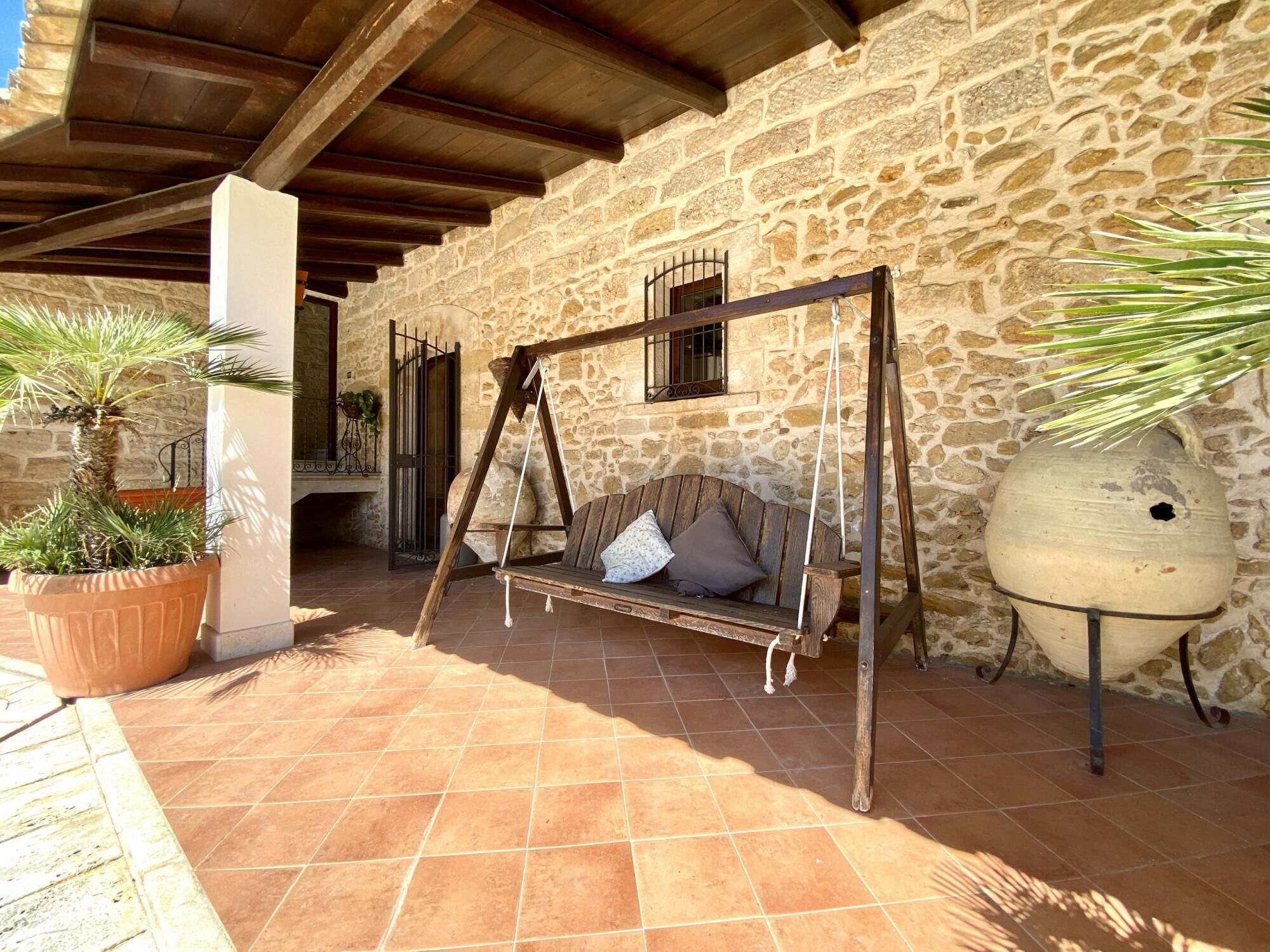 Une terrasse aux murs de pierre, agrémentée d’une balançoire en bois, de plantes en pot et d’un mobilier extérieur rustique.