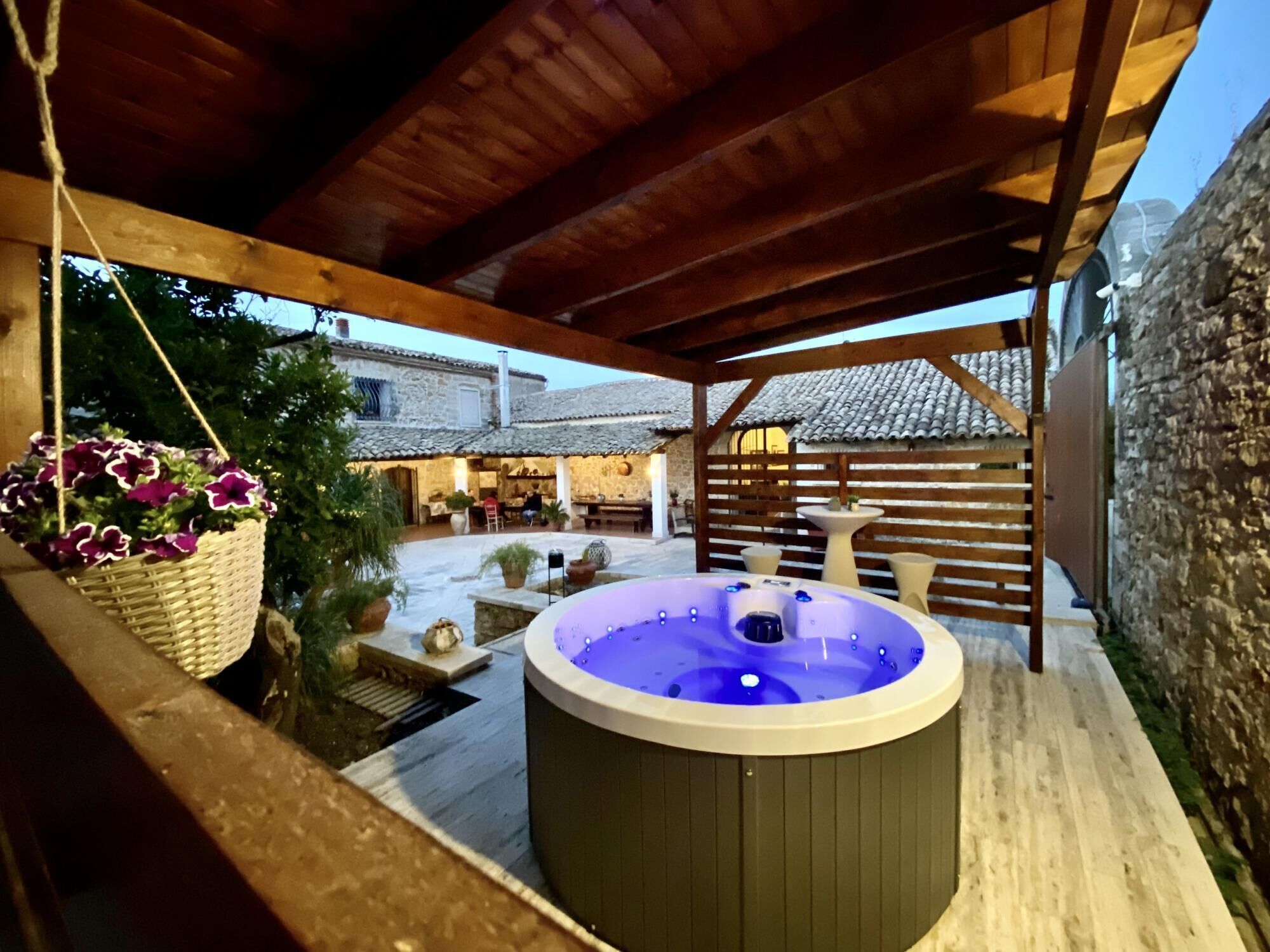 Un jacuzzi bajo una pérgola de madera con paredes de piedra y un edificio de piedra al fondo.