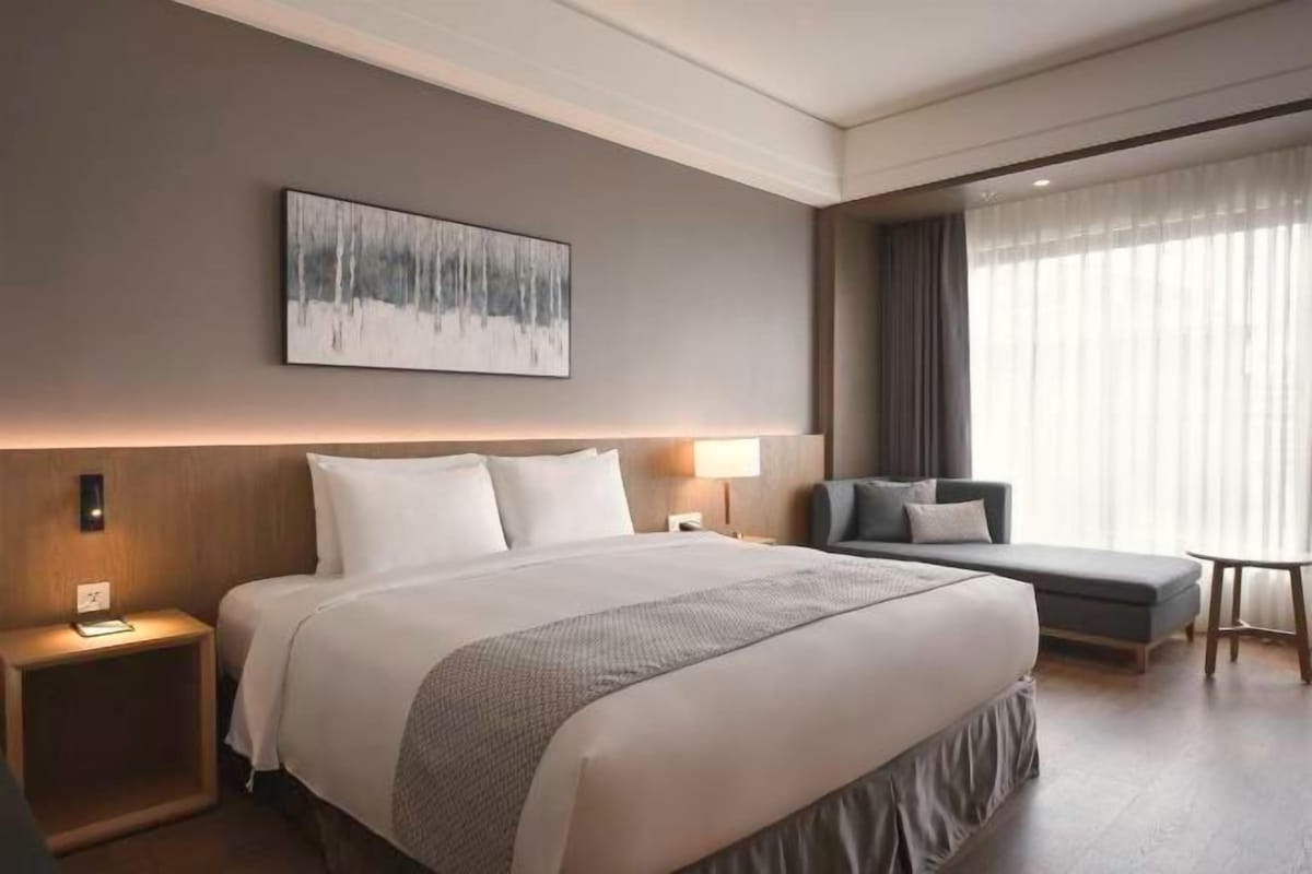 A modern hotel room with a large bed, bedside tables, a desk, a chair, a large window with curtains, and a painting on the wall.
