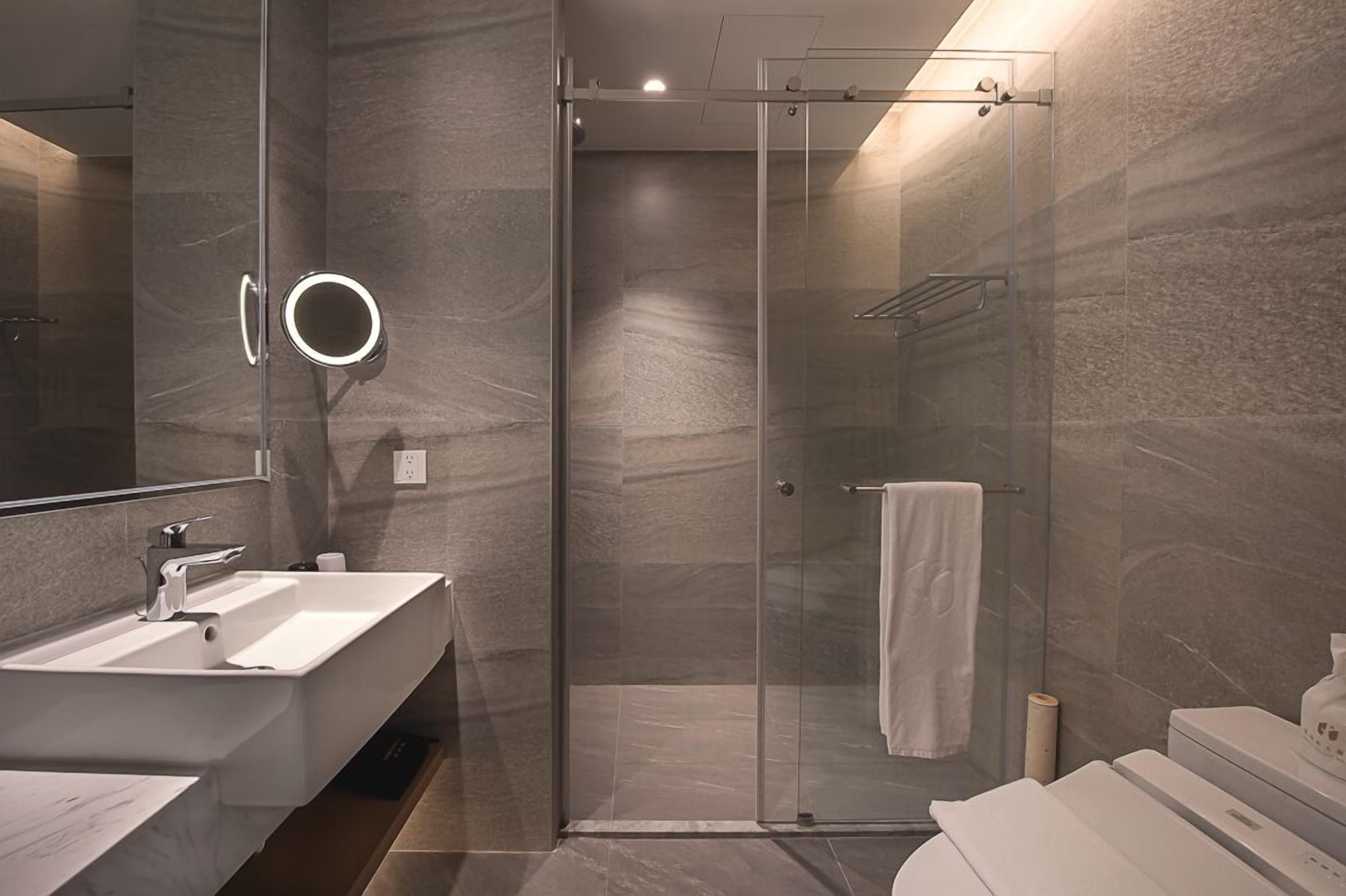 Un baño moderno con ducha envuelta en vidrio, un espejo grande con luz y un lavamanos blanco.
