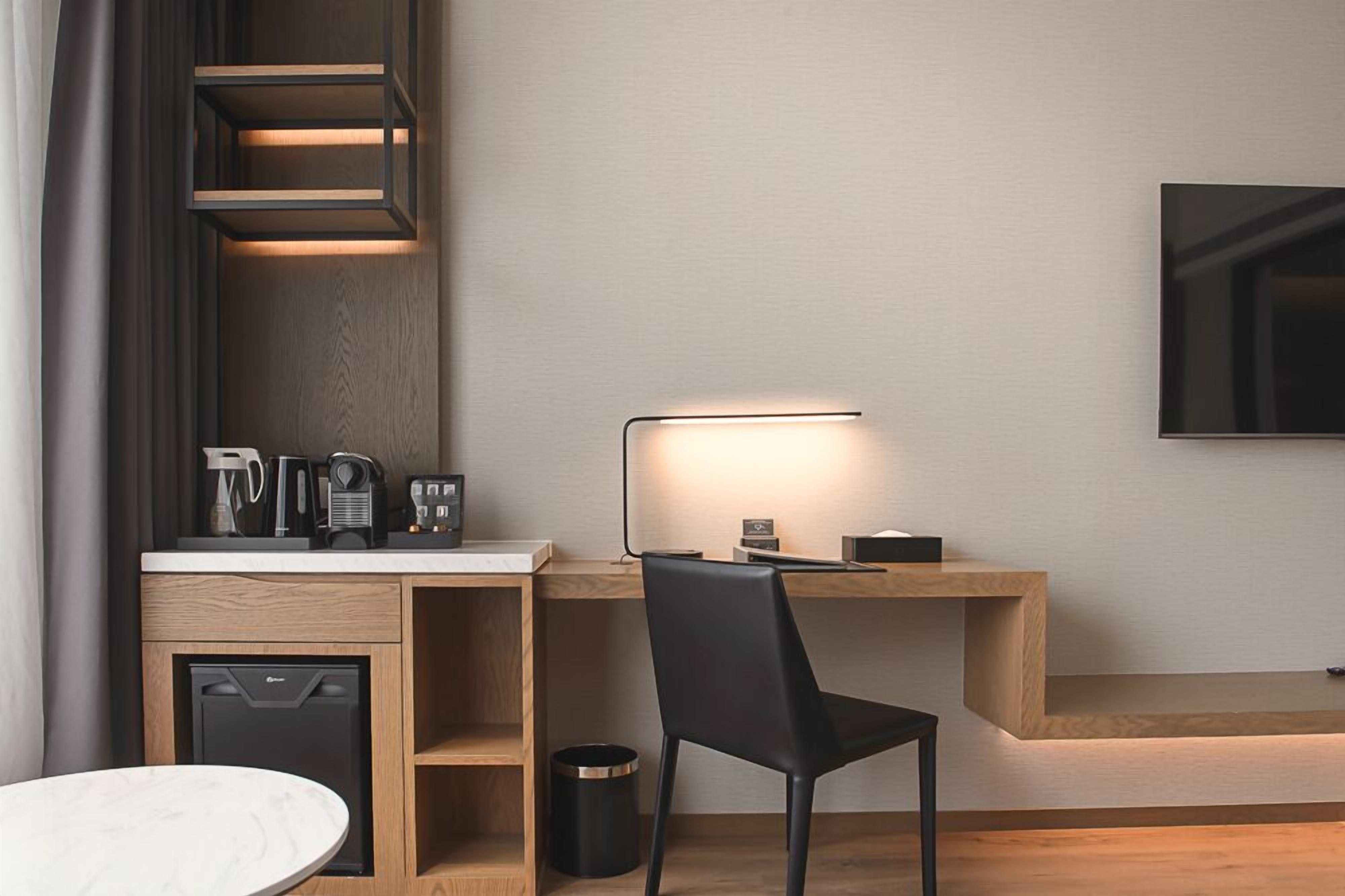 A modern hotel room with a wooden desk, a black chair, a small coffee machine, a television mounted on the wall, and a compact dishwasher.