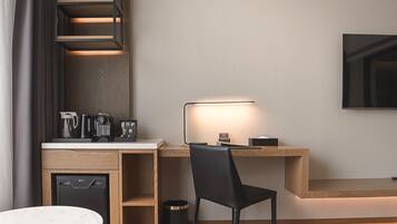 A modern hotel room with a wooden desk, a black chair, a small coffee machine, a television mounted on the wall, and a compact dishwasher.