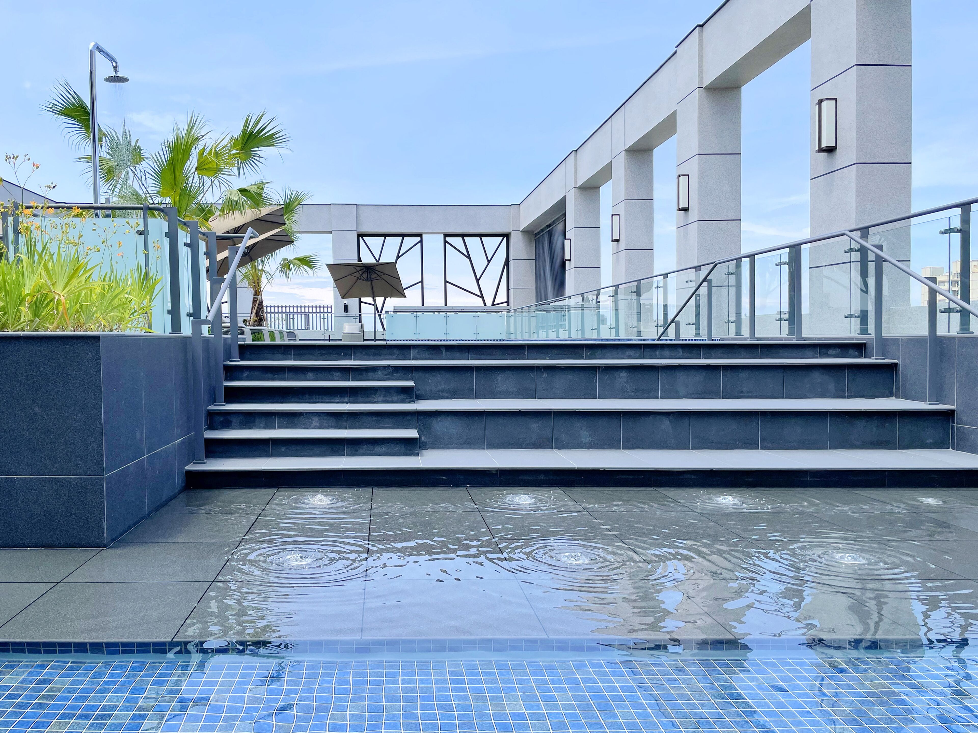 Un área de piscina moderna al aire libre con piso de baldosas azules brillantes, una escalera que conduce a una zona de estar cubierta y una barandilla de vidrio con vista a la piscina.