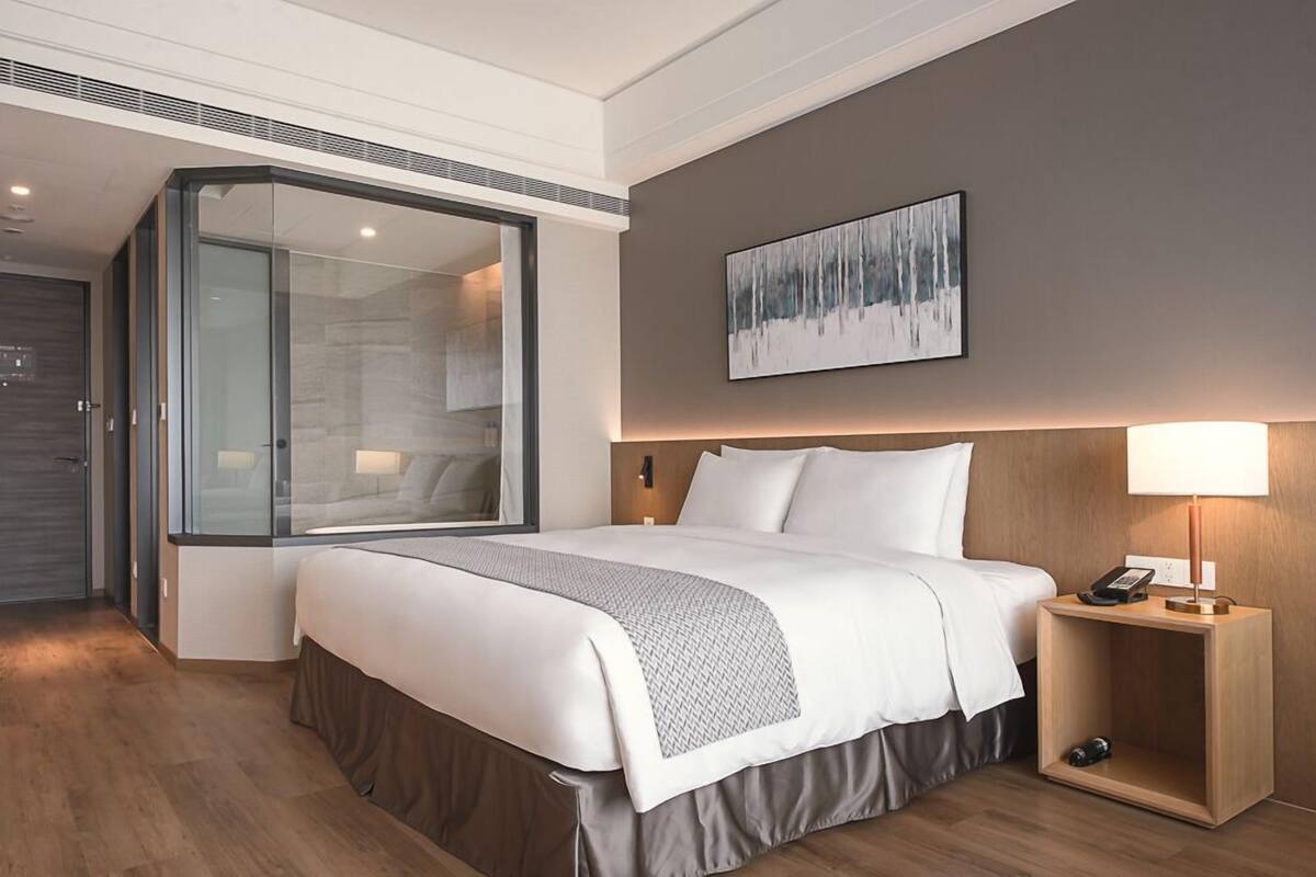 A modern hotel room with a large bed, a glass partition, a wooden nightstand, and a painting on the wall.