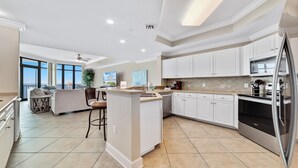 A modern kitchen with white cabinets, a stainless steel refrigerator, and a large island. There is a dining area with a sofa and chairs, and a large window offering a view. - New Owner, New to the Rental Market, Lazy River/Slide!! (Orange Beach)