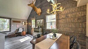 A rustic dining area with a wooden table, chairs, and a chandelier. A deer head is mounted on the wall above the table.