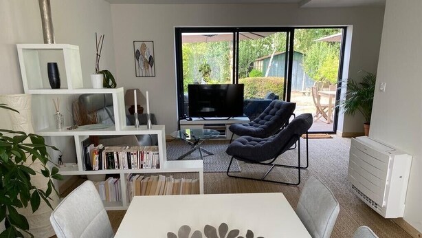 Un salon moderne avec une table blanche, des fauteuils gris, un canapé noir, une bibliothèque et un téléviseur.