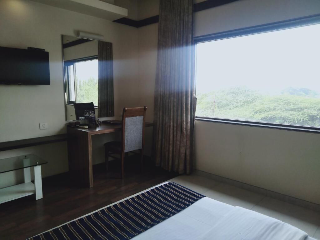 Ein Hotelzimmer mit Bett, Schreibtisch, Stuhl, einem an der Wand befestigten Fernseher und einem großen Fenster mit Blick auf Grünflächen.