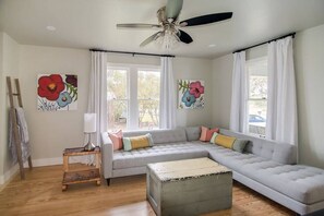 A living room with a grey sectional sofa, a small wooden table, a ceiling fan, and two windows with white curtains. - Pet-Friendly Craftsman Home in Fredericksburg (Fredericksburg)