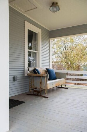 A porch with a wooden bench and cushions, a window, and a view of trees.