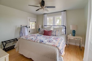 A bedroom with a bed, a ceiling fan, a window with curtains, a bedside table with a lamp, and a wooden floor.