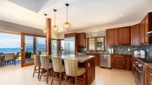 A modern kitchen with wooden cabinets, a stainless steel refrigerator, and a large island with bar stools. - Breathtaking Oceanfront Puamana Retreat – 5 Bedrooms, Modern & Spacious (Lahaina)