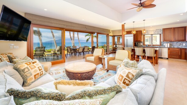 A spacious living area with a large sofa, a round coffee table, and a view of the ocean through large windows. There are ceiling fans, a flat-screen TV, and a kitchen area visible. - Breathtaking Oceanfront Puamana Retreat – 5 Bedrooms, Modern & Spacious (Lahaina)