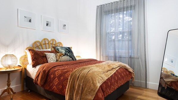 Un dormitorio ordenado con cama, mesita de noche, espejo y ventana con cortinas translúcidas.