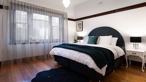 Ein Schlafzimmer mit Bett, Nachttisch, Lampe, einem Fenster mit durchscheinenden Vorhängen und einem gerahmten Bild an der Wand.