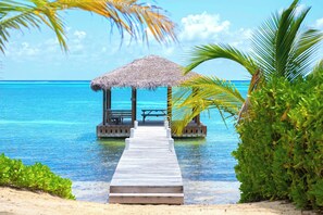 A wooden pier leading to a thatched-roof structure with a bench and table, overlooking a clear blue sea.