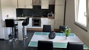 A modern kitchen with white cabinets, a stove, oven, microwave, and a sink. There is a dining area with a table and chairs, and a window providing natural light. - Beautiful holiday apartment on the Moselle (Piesport)