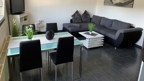 A modern living room with a grey sofa, a glass dining table, black chairs, a flat-screen TV on a white stand, and a painting on the wall. - Beautiful holiday apartment on the Moselle (Piesport)