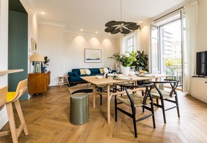 Un salon moderne avec un parquet en bois disposé en chevrons, une table à manger avec des chaises, un canapé avec des coussins, un téléviseur et une grande fenêtre donnant sur des immeubles.
