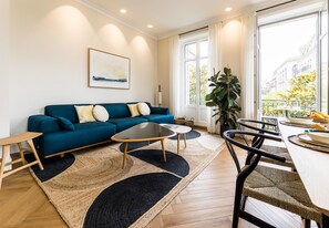 Un salon moderne avec un canapé bleu, deux tables basses, une table à manger avec des chaises et une grande fenêtre donnant sur des immeubles et des arbres.