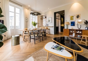 Un salon moderne comprenant un coin repas, un plancher en bois, une table basse en verre et une grande fenêtre avec des rideaux.