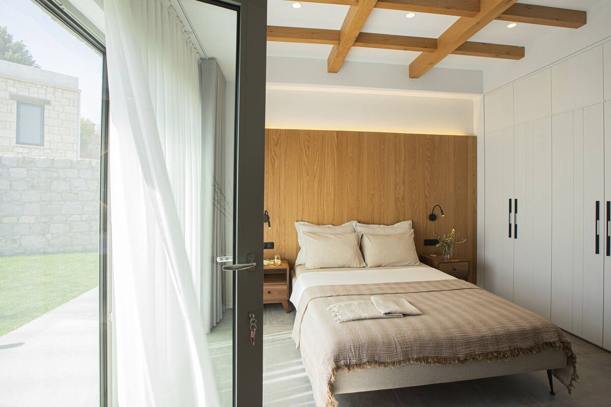 A modern bedroom with a large bed, wooden ceiling beams, a nightstand with a lamp, and a view of a stone building outside.