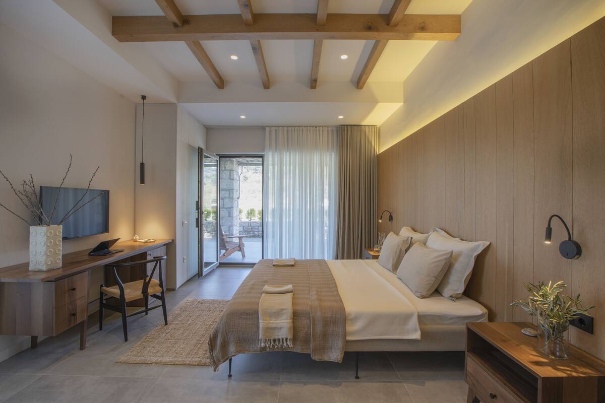 A modern bedroom with a large bed, a desk, and a chair. There is a TV on the desk and a window with curtains.