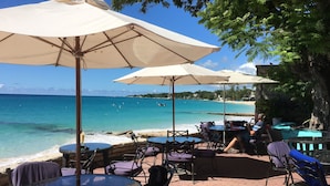 A beachside café with outdoor seating, including tables and chairs, under large umbrellas, overlooking the ocean.