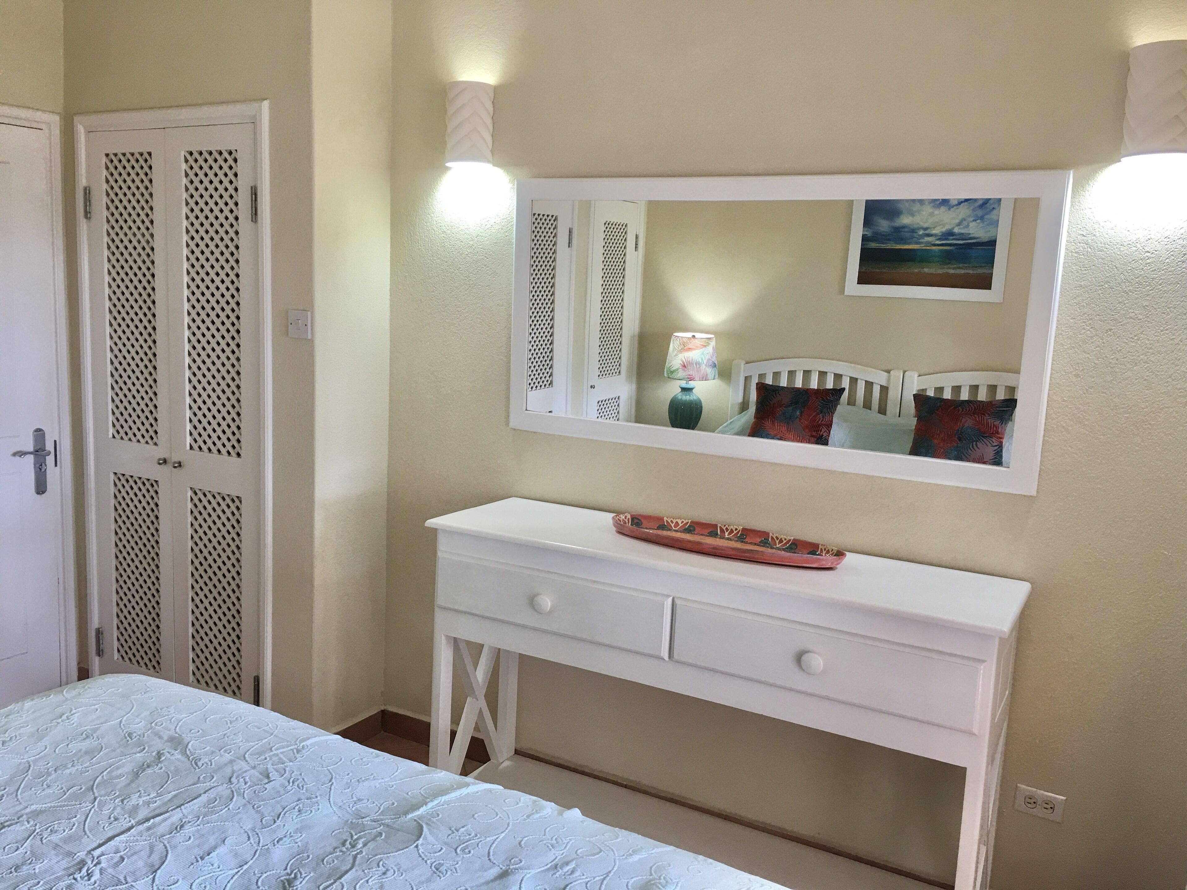 Une chambre à coucher avec un lit, une commode blanche, un miroir et une porte ornée d’un motif en treillis.