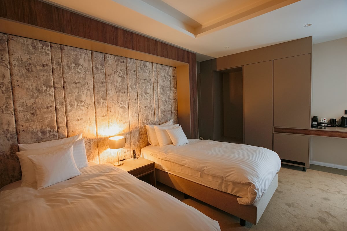 A hotel room with two beds, a wooden headboard, a bedside table with a lamp, and a kitchenette area.