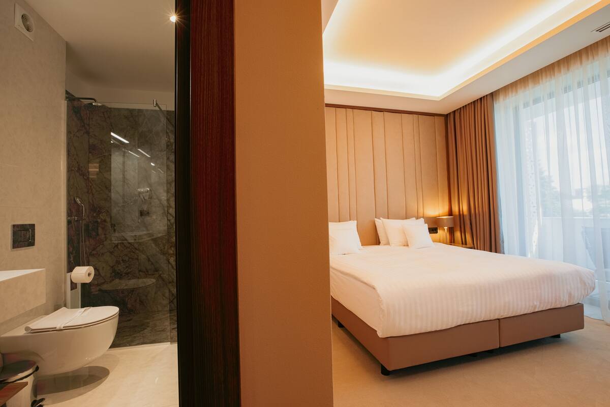 A modern hotel room with a marble shower, a large bed, and a view of the city through sheer curtains.