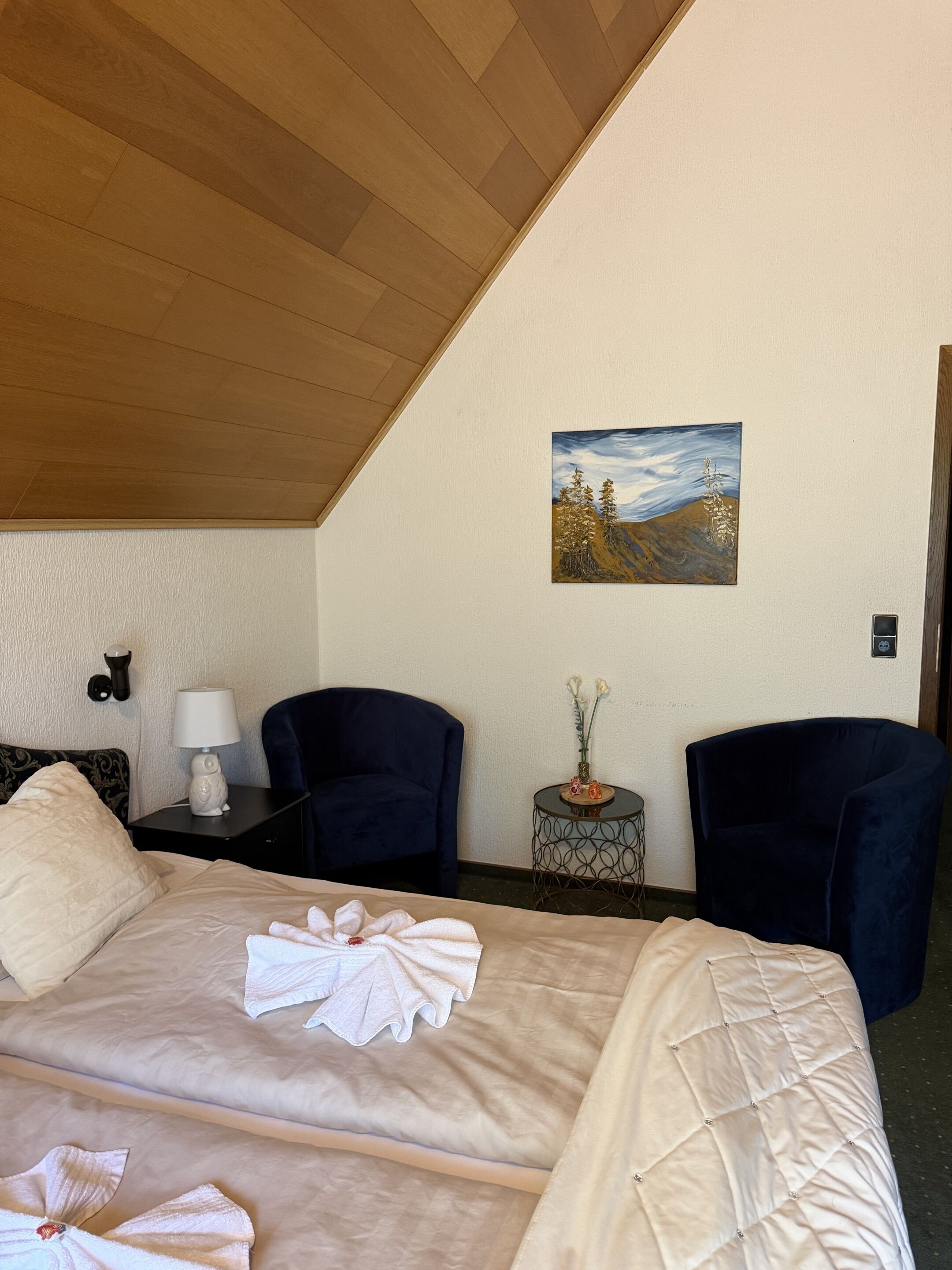 A hotel room with a bed, two chairs, a painting, a small table with a vase, and a lamp.