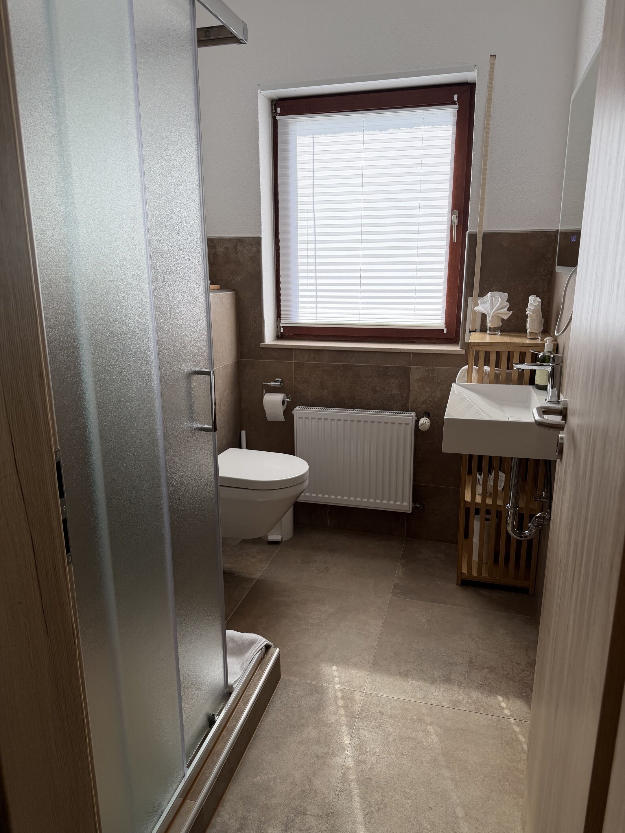 A bathroom with a toilet, a radiator, a window with blinds, a sink with a mirror, and a shower area.