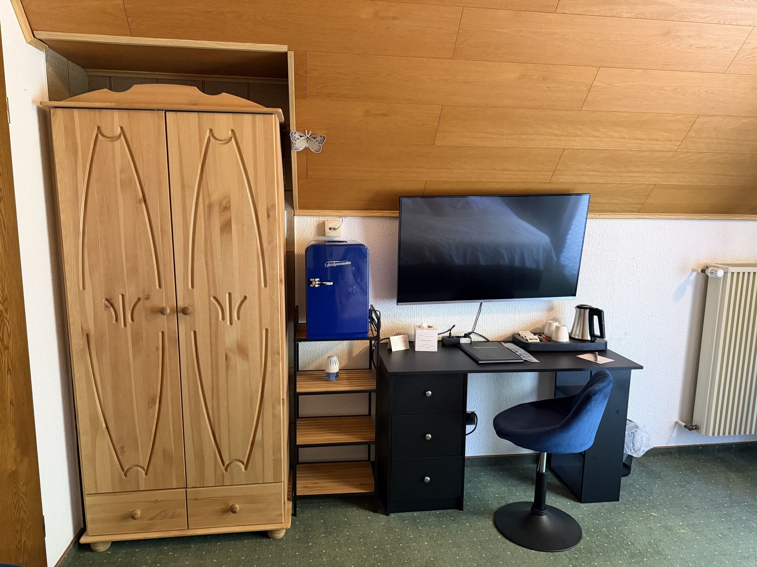 Una habitación con un armario de madera, un escritorio con silla, un dispensador de agua azul, un televisor de pantalla plana y un radiador.