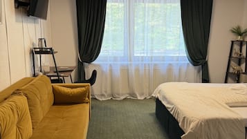 A room with a sofa, a bed, a TV mounted on the wall, and a window with curtains.