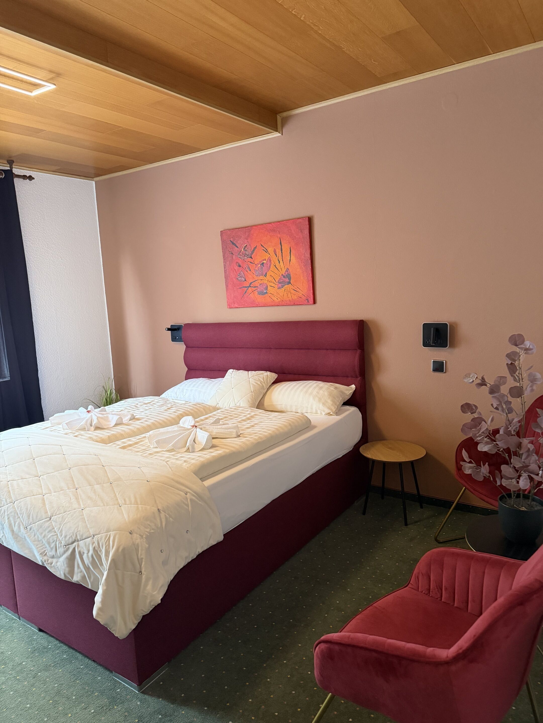 A bedroom with a bed, a red chair, a small stool, a painting on the wall, and a potted plant.