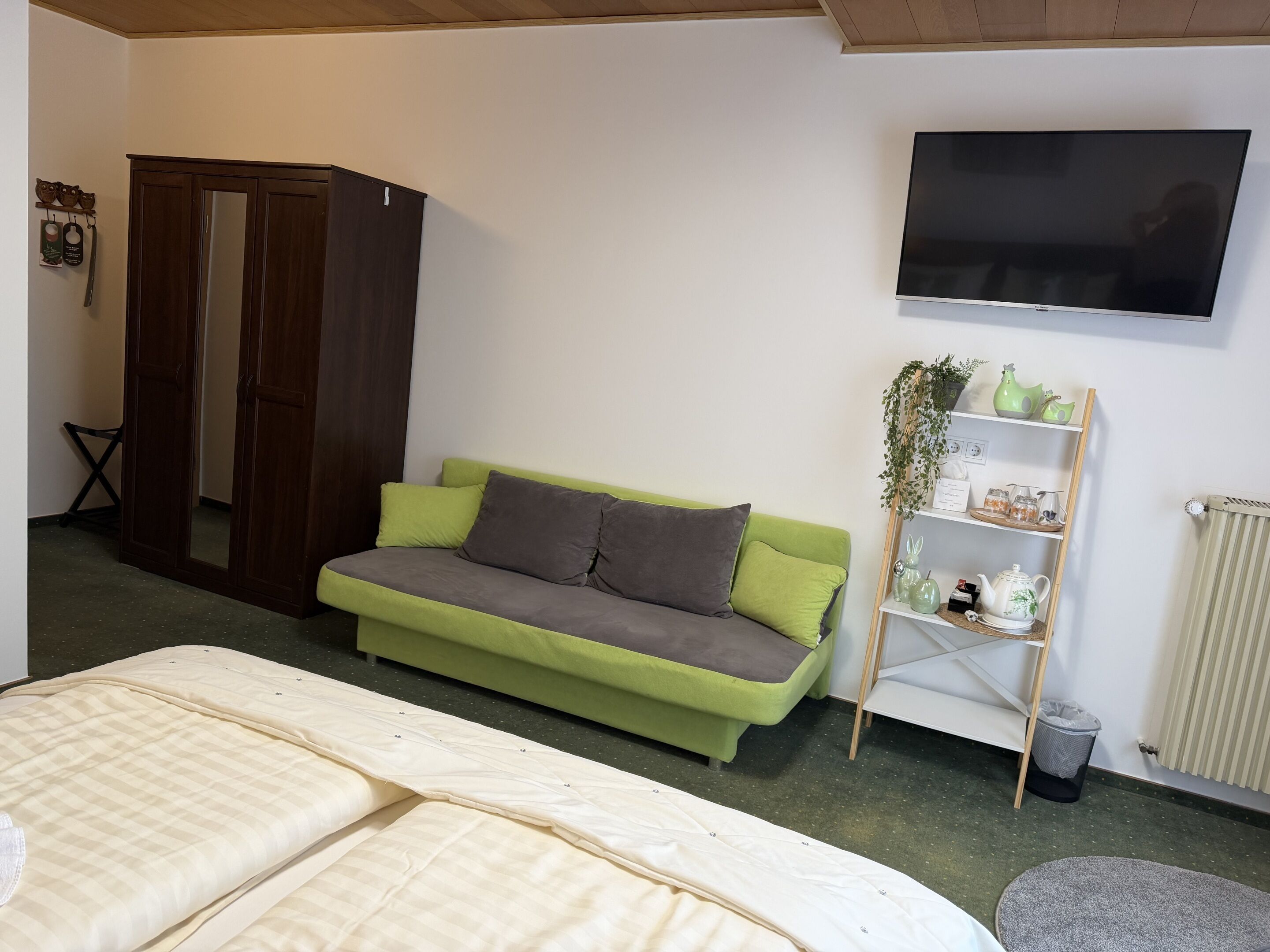 A room with a green sofa, a bed with a striped cover, a wooden wardrobe, a TV on the wall, and a shelf with decorative items.
