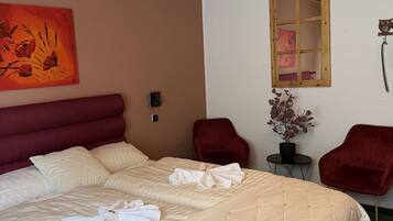 Habitación de hotel con una cama grande, dos sillas rojas, una mesita con una planta, un cuadro en la pared y una ventana con marco de madera.