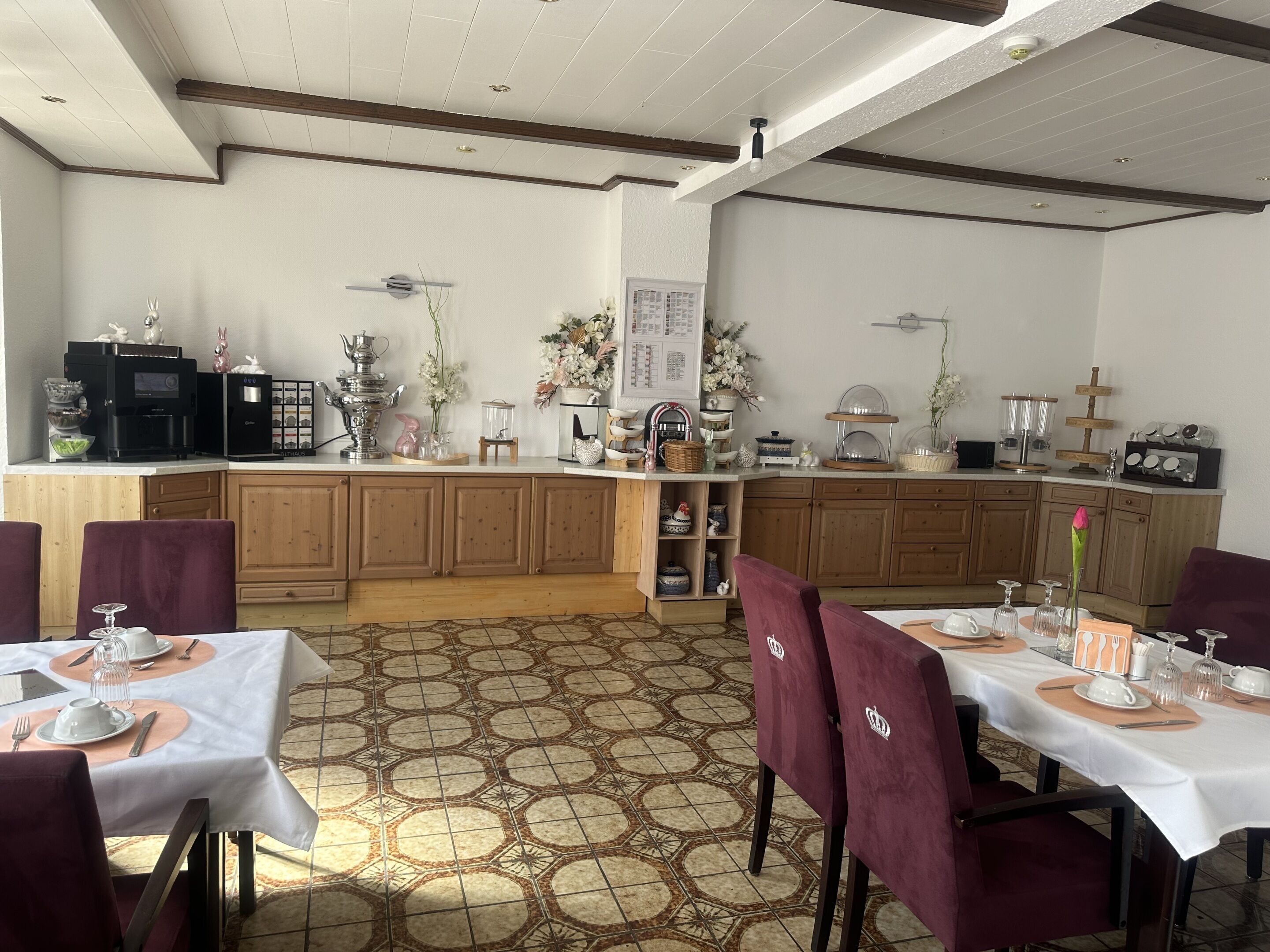 A dining area with a kitchen in the background, featuring wooden cabinets, a tiled floor, and a white ceiling with wooden beams.
