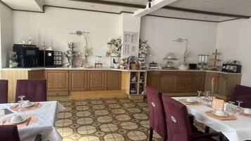 A dining area with a kitchen in the background, featuring wooden cabinets, a tiled floor, and a white ceiling with wooden beams.