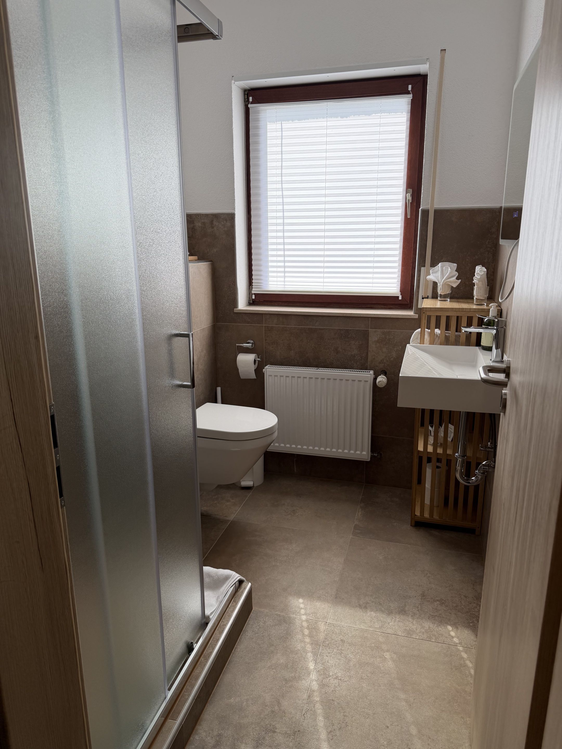 Une salle de bain comprenant des toilettes, un radiateur, une fenêtre avec stores, un lavabo avec un miroir et un espace douche.