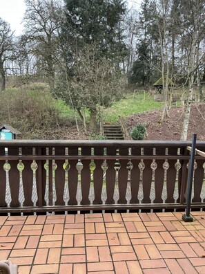 A brick patio with a decorative railing, a small wooden bench, and a view of a wooded area with a stone structure in the background. - Hotel Eulenhof (Gransdorf)