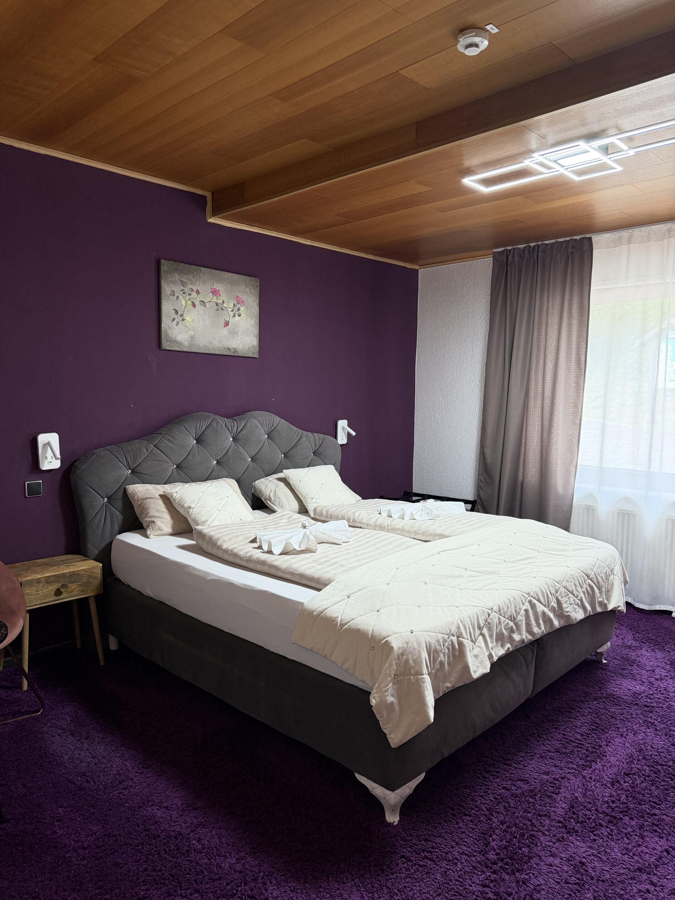 A bedroom with a bed, purple walls, a ceiling light fixture, and a window with curtains.