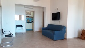 Una sala de estar con un sofá azul, un televisor montado en la pared y un área de cocina visible a través de un umbral abierto.